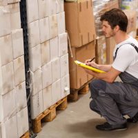 Side view of worker crouching down, looking at goods and writing something in yellow clipboard. Man wearing uniform and white t shirt. Background of warehouse with lots of boxes.